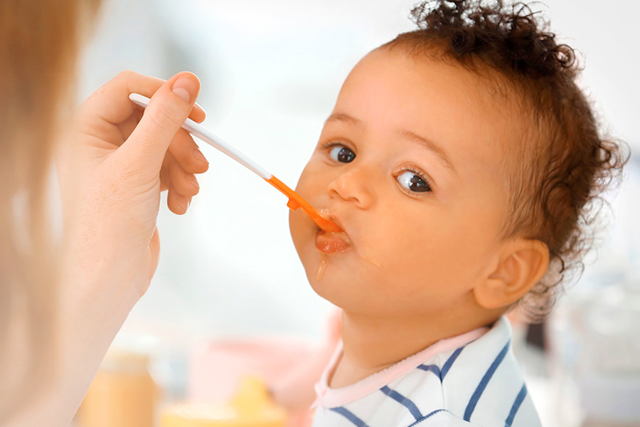 Bebê sendo alimentado na boca por uma mulher com uso de uma colher branca e laranja