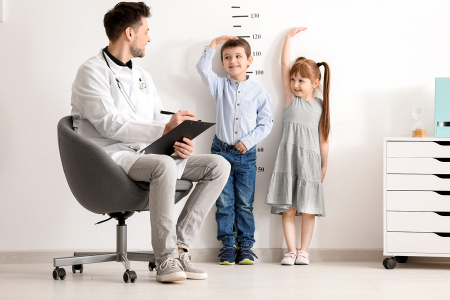 Médico com uma prancheta sentado em uma cadeira medindo a altura de duas crianças por meio de marcações na parede