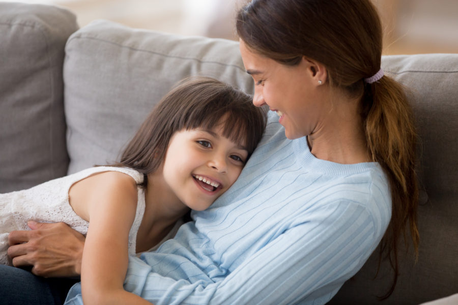 Mãe e filha conversando e se abraçando felizes no sofá com o objetivo de prevenir o estresse tóxico