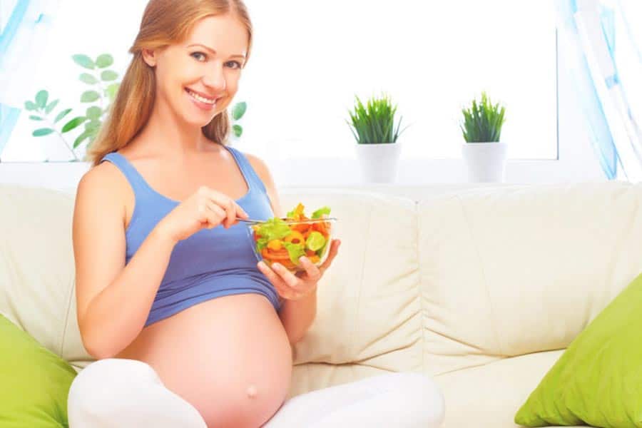 Mulher grávida sentada no sofá comendo uma salada. Essa é uma das formas de aliviar os enjoos na gravidez