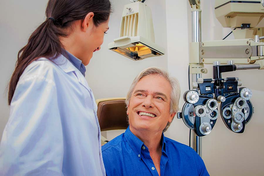 Paciente sentado em uma cadeira no consultório, durante uma consulta oftalmológica, olhando sorridente para a oftalmologista.
