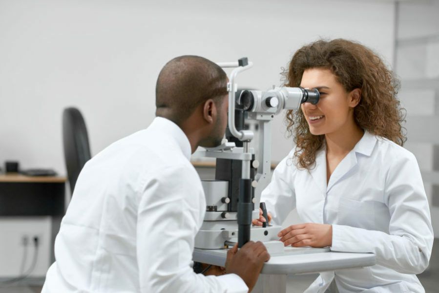 Homem negro tendo o olho examinado por uma oftalmologista negra. Representação dos sinais e sintomas de alerta que indicam que é preciso procurar um oftalmologista.