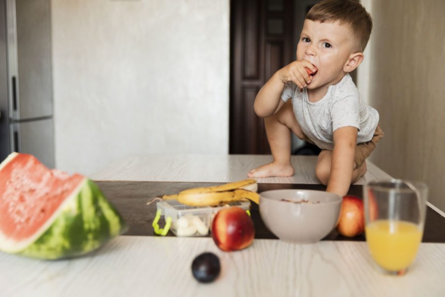Menino hiperativo com dificuldade alimentar proveniente do TDAH em cima de uma mesa repleta de alimentos saudáveis