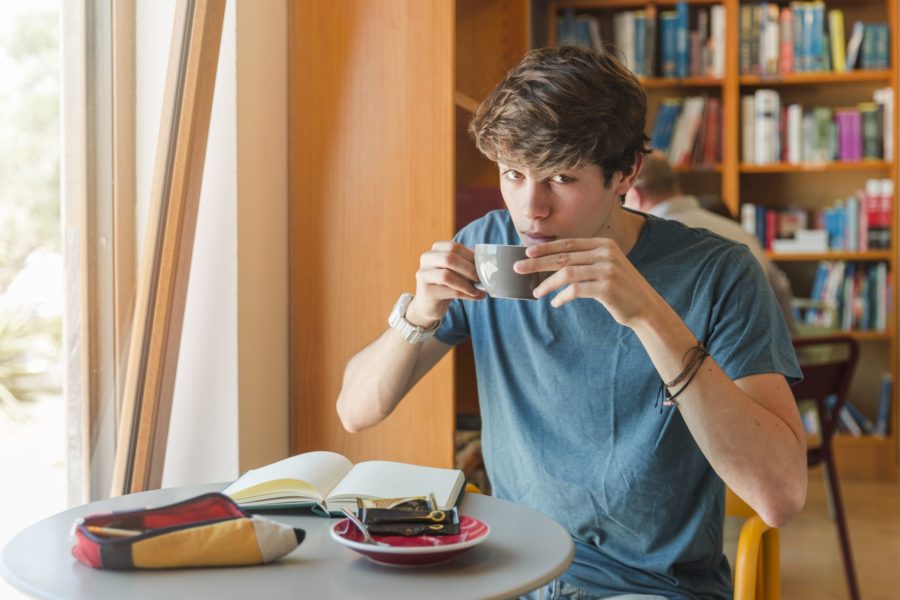 Adolescente tomando uma xícara de café enquanto está em uma biblioteca estudando com seu estojo e caderno em cima da mesa