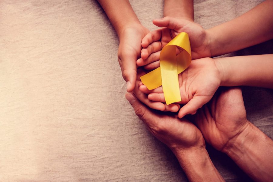 Mãos de adulto e criança segurando uma fita amarela de câncer sobre um fundo bege. O intuito é alertar sobre os tipos de câncer infantil mais comuns