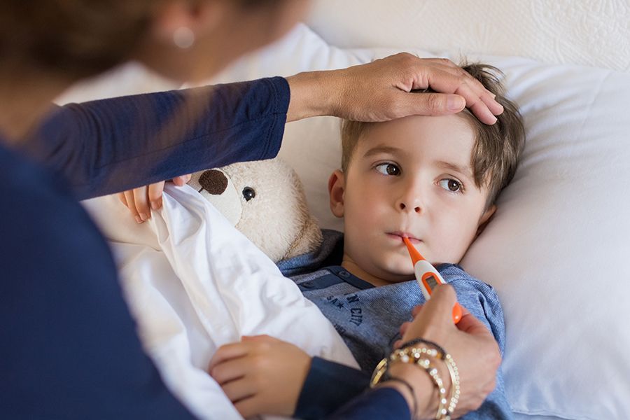 vista de cima de menino deitado na cama com seu ursinho de pelúcia e sua mãe medindo a temperatura com um termômetro na boca dele