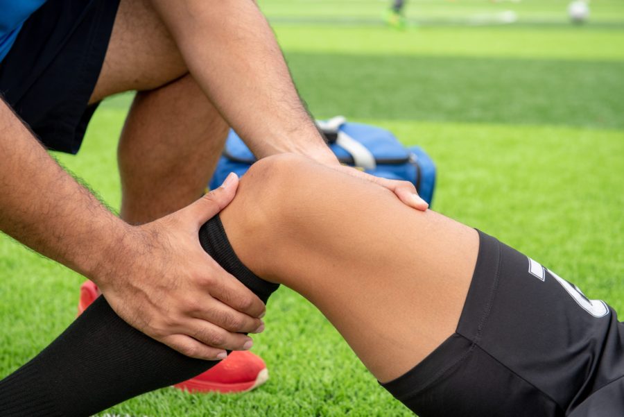 Jogador de futebol dentro do campo tendo o joelho examinado por um fisioterapeuta por causa de uma lesão que sofreu durante uma partida. Existe a possibilidade de ser ruptura do menisco.