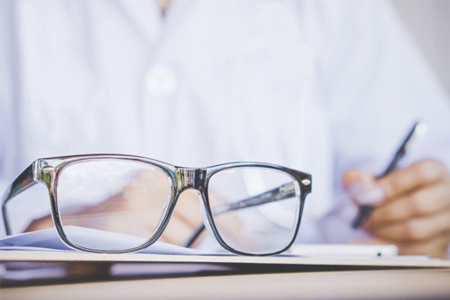 Óculos em primeiro plano e, desfocado, em segundo plano médico oftalmologista escreve uma receita de óculos para um paciente