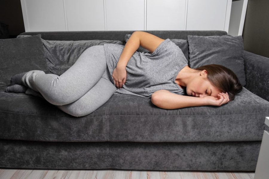 Adolescente deitada de olhos fechados em um sofá com um das mãos na barriga e outra no rosto indicando que está sofrendo de alguma dor proveniente de doença inflamatória intestinal