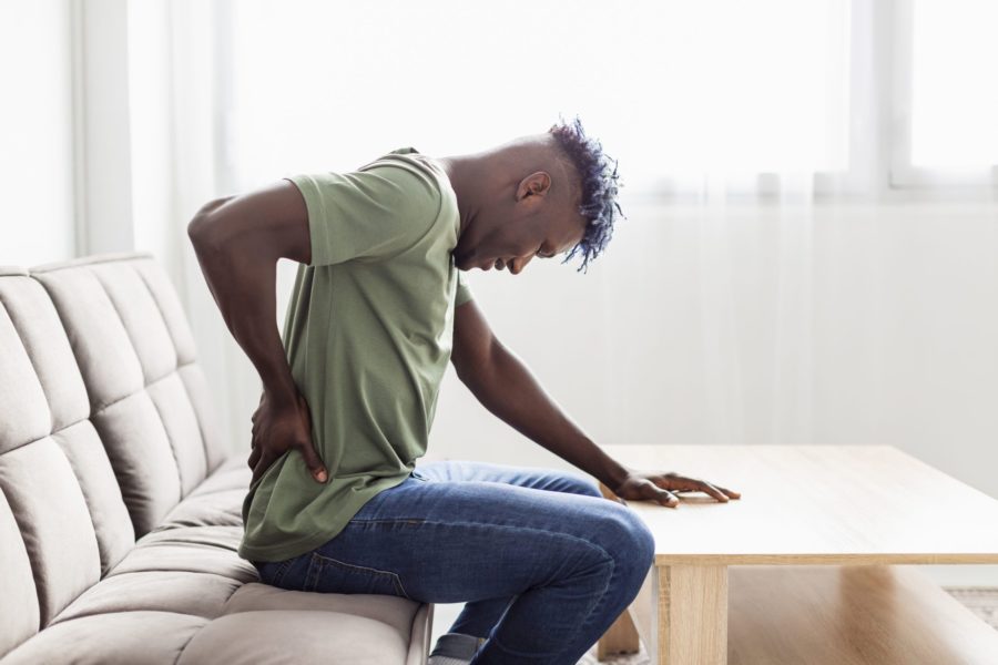 Homem sentado no sofá com uma das mõas na parte inferior das costas, a cabeça abaixada e a outra mão apoiada na mesa. Ele está com dor ciática