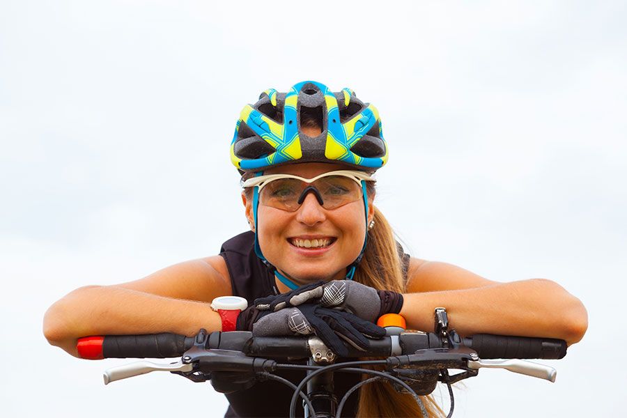 Mulher sorridente debruçada no guidão da bicicleta usando capacete, óculos esportivos, luvas e um relógio para medir os batimentos cardíacos