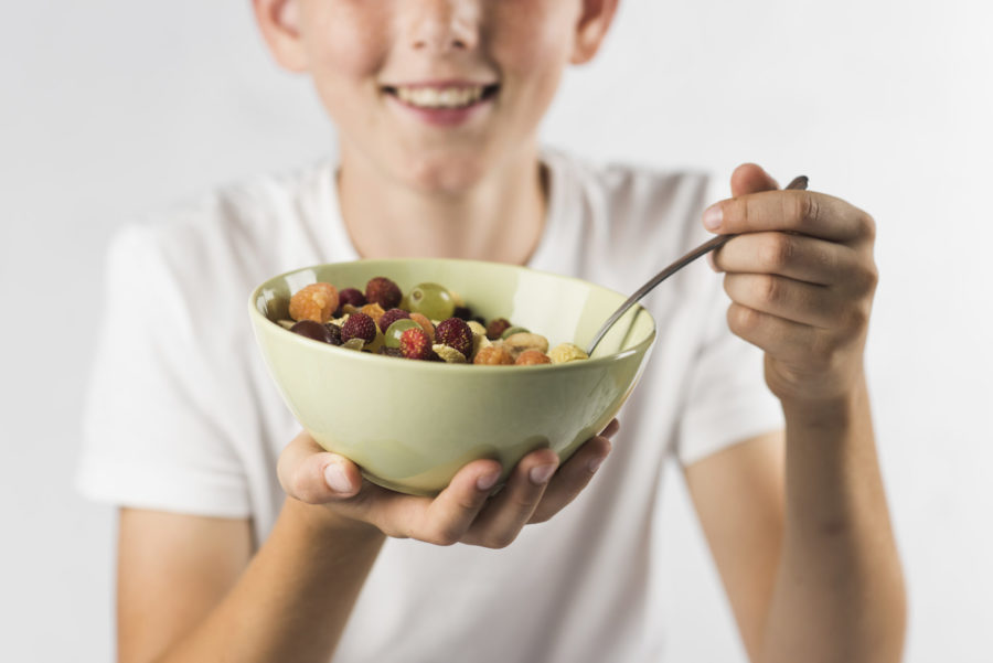 Sobre um fundo branco, retrato de um garoto sorridente segurando uma tigela de salada de fruta. Representação do veganismo na infância e na adolescência.