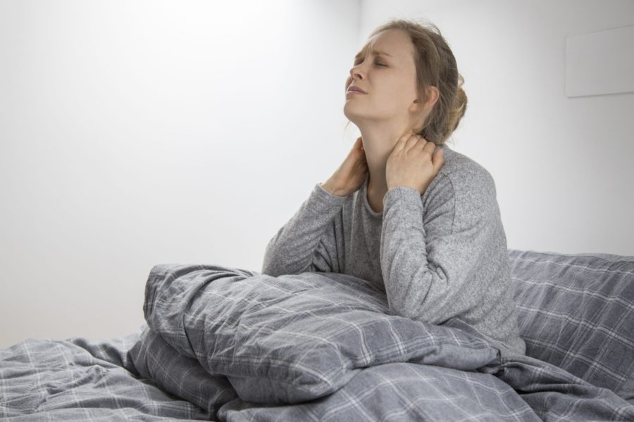 Mulher sentada na cama com as mãos na parte de trás do pescoço e a cara com feições que denotam que ela está sentindo dor por conta da fibromialgia
