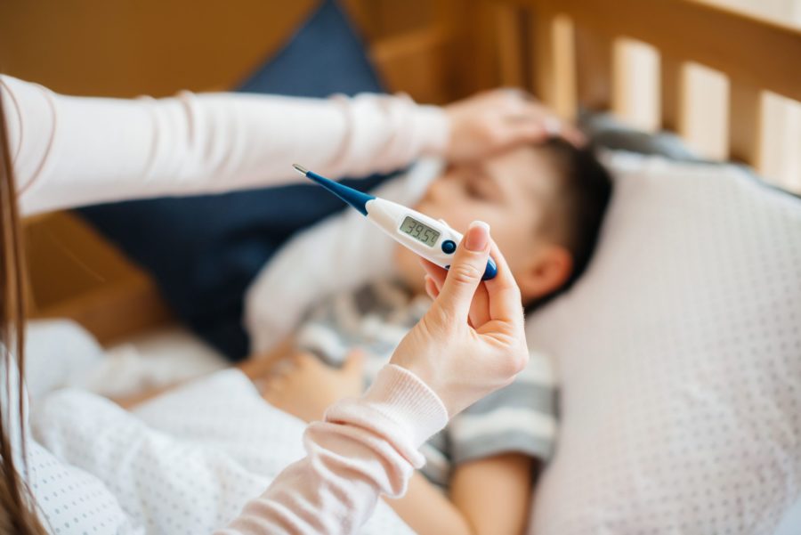 Mão medindo a temperatura do seu filho com um termômetro digital. Está marcando 39,5 graus e a mãe está encostando a mão na testa do menino que está deitado na cama.