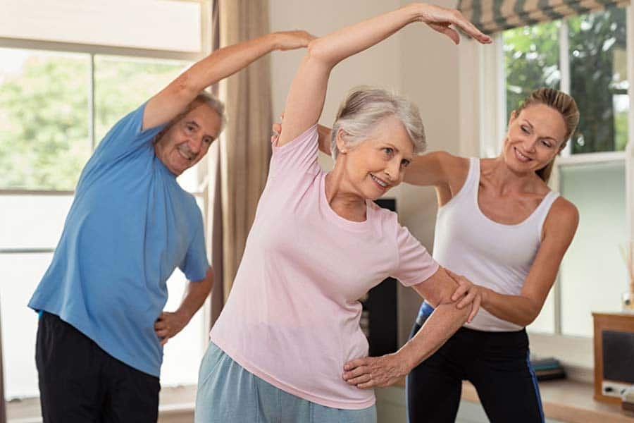 Para prevenir ou tratar a osteoporose, dois idosos fazendo exercício de alongamento com auxílio de uma instrutora mais jovem.