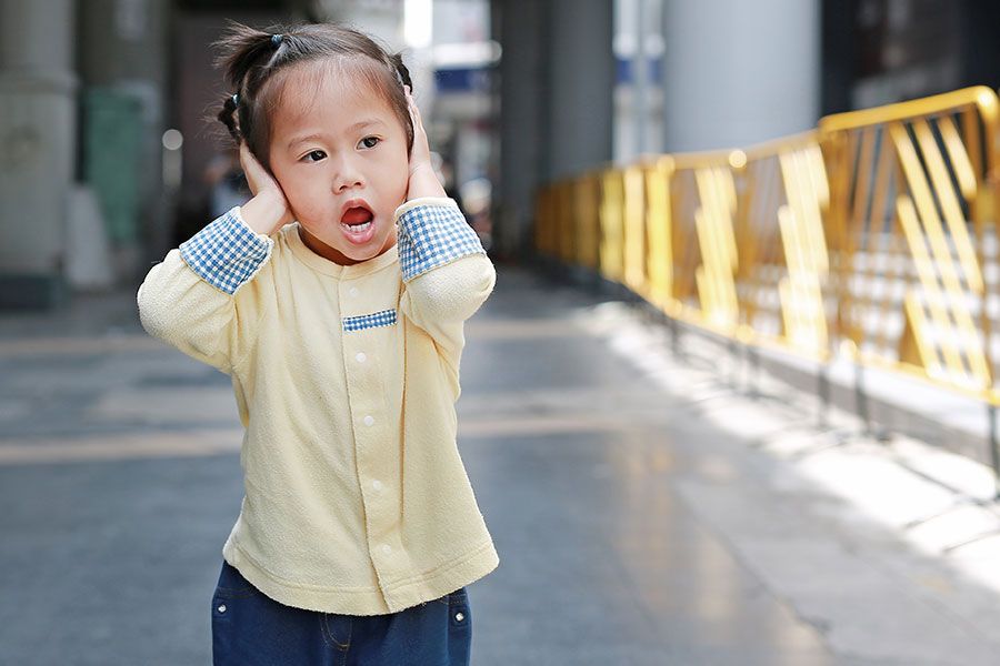 Em ambiente externo, menina asiática com autismo tapando as orelhas com as mão e com um expressão de desconforto.
