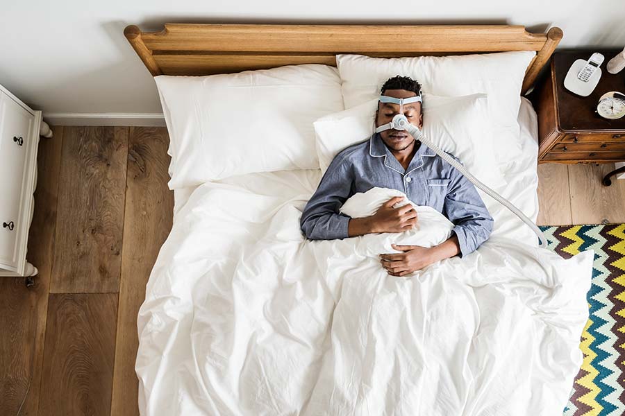 Vista de cima de um homem com uma máscara para tratamento da apneia do sono deitado na cama no seu quarto.