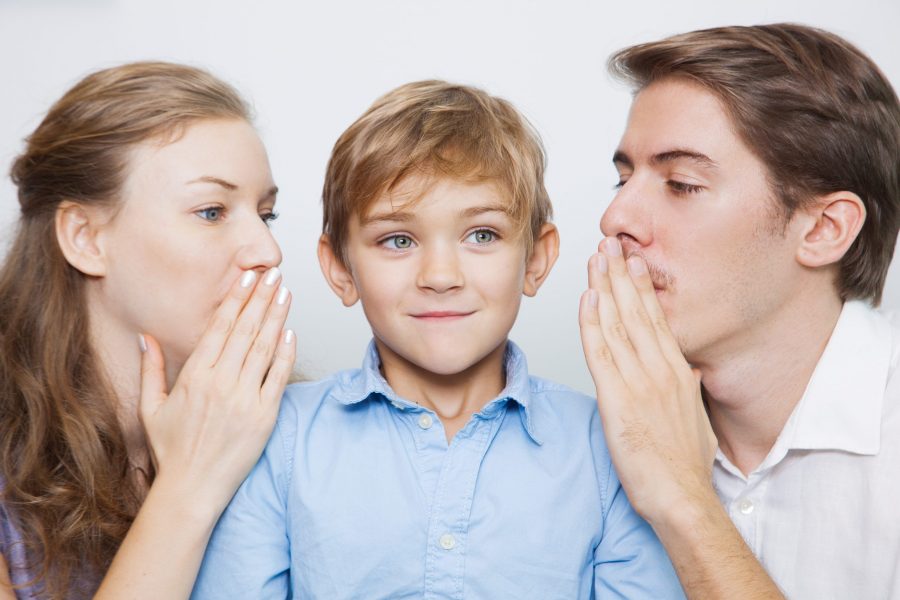 Pai e mãe de uma criança autista falando algo no ouvido dele. Representação das 5 principais coisas para nunca dizer a um autista.