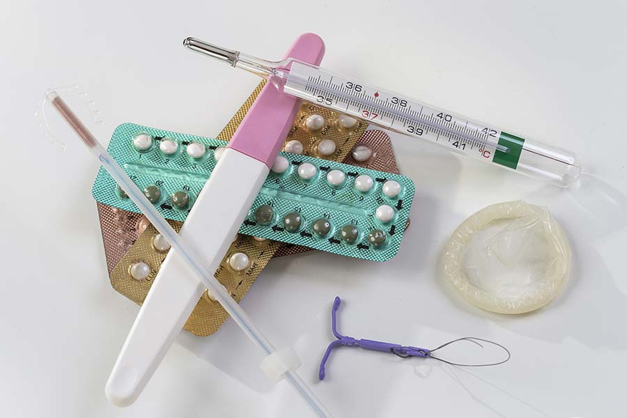 Vários métodos contraceptivos (diu, camisinha, pílula, teste de gravidez, injeção) e um termômetro sobre um fundo branco.