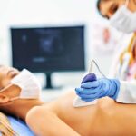 Médica de máscara realizando um exame de mamotomia guiada por ultrassom em uma paciente que também está de máscara.
